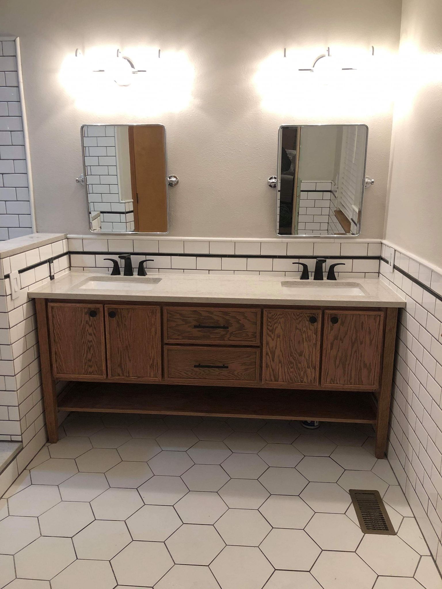Bathroom with a wooden double vanity, black fixtures, mirrors, and hexagonal white tile.
