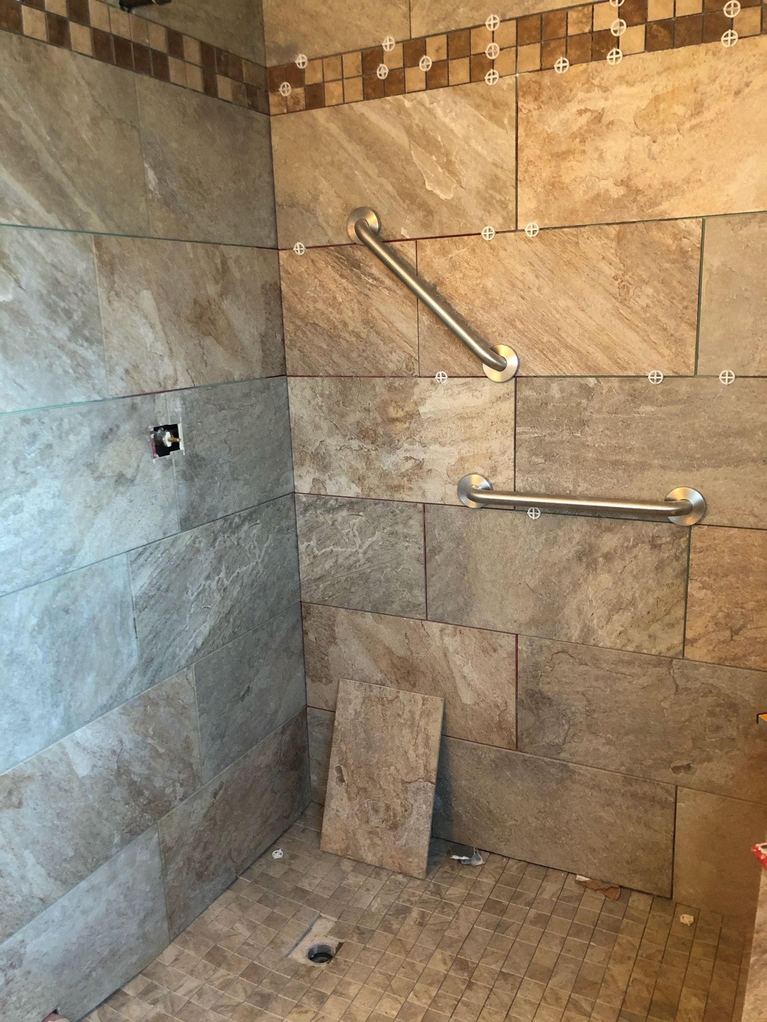 Shower stall under construction, featuring tan stone tiles and two silver grab bars installed on the wall.
