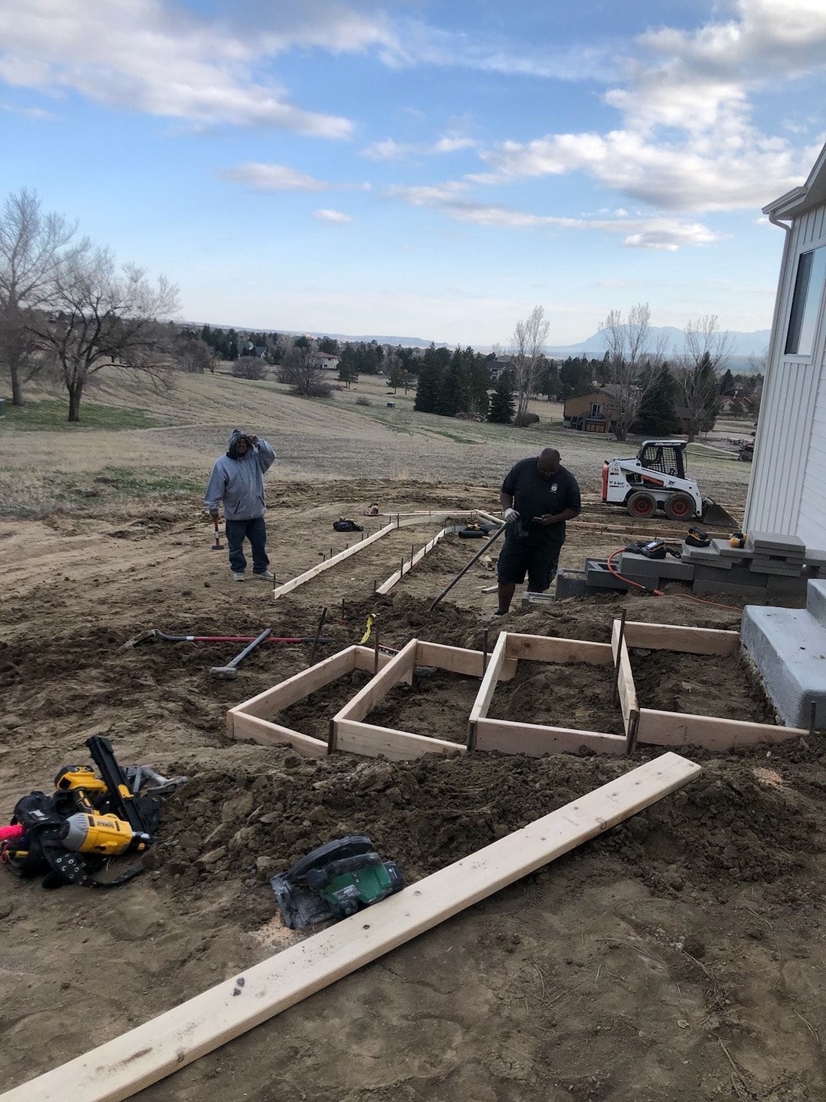Two people constructing forms for concrete on a dirt lot, near a building. Tools and materials are scattered around.