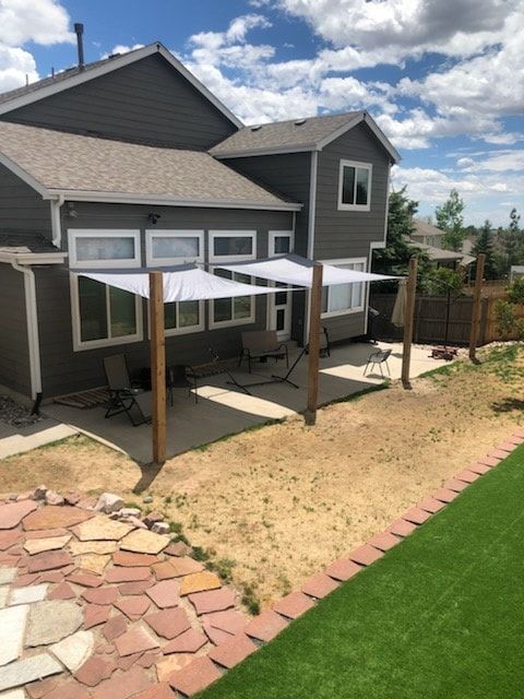 Backyard with gray house, concrete patio, two white sun sails on wooden posts, and patchy lawn.