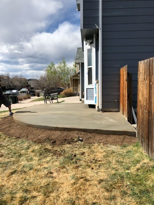 A newly poured concrete patio next to a blue house with a brown wooden fence, and a grassy yard.