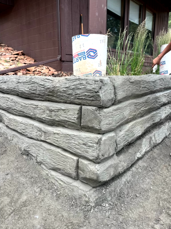 Corner of a concrete retaining wall designed to look like stacked logs; brown building background.