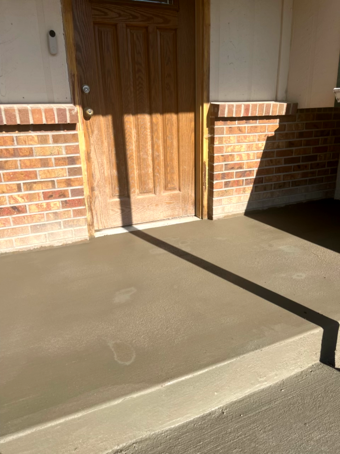 Freshly poured concrete front porch with a step, next to a brick wall and wooden door.