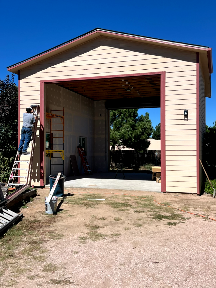 Man on a ladder working on the exterior of a tan building with open garage, clear blue sky.