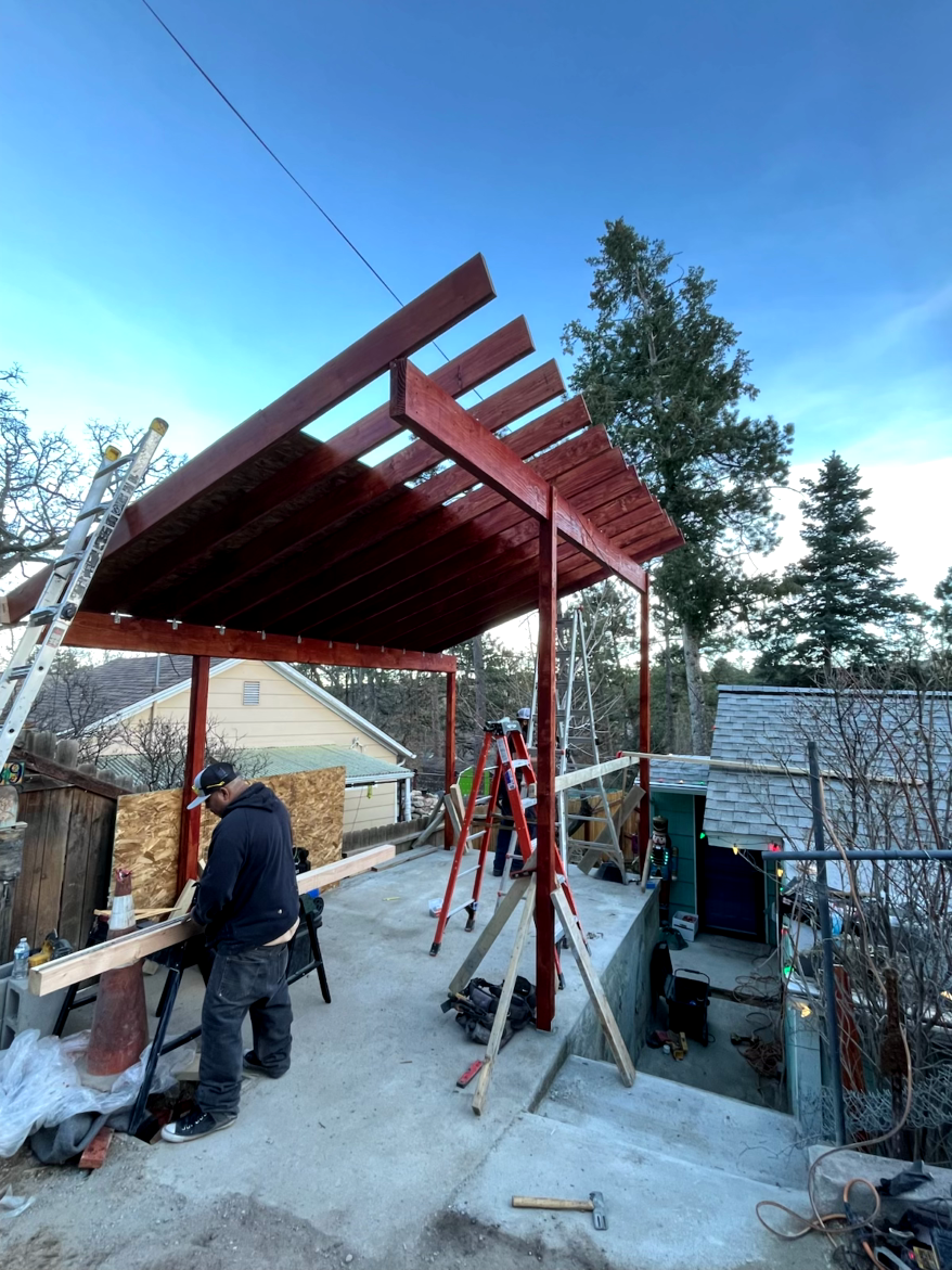 Construction of a red-painted pergola outside a house. Workers use tools on concrete. Blue sky overhead.