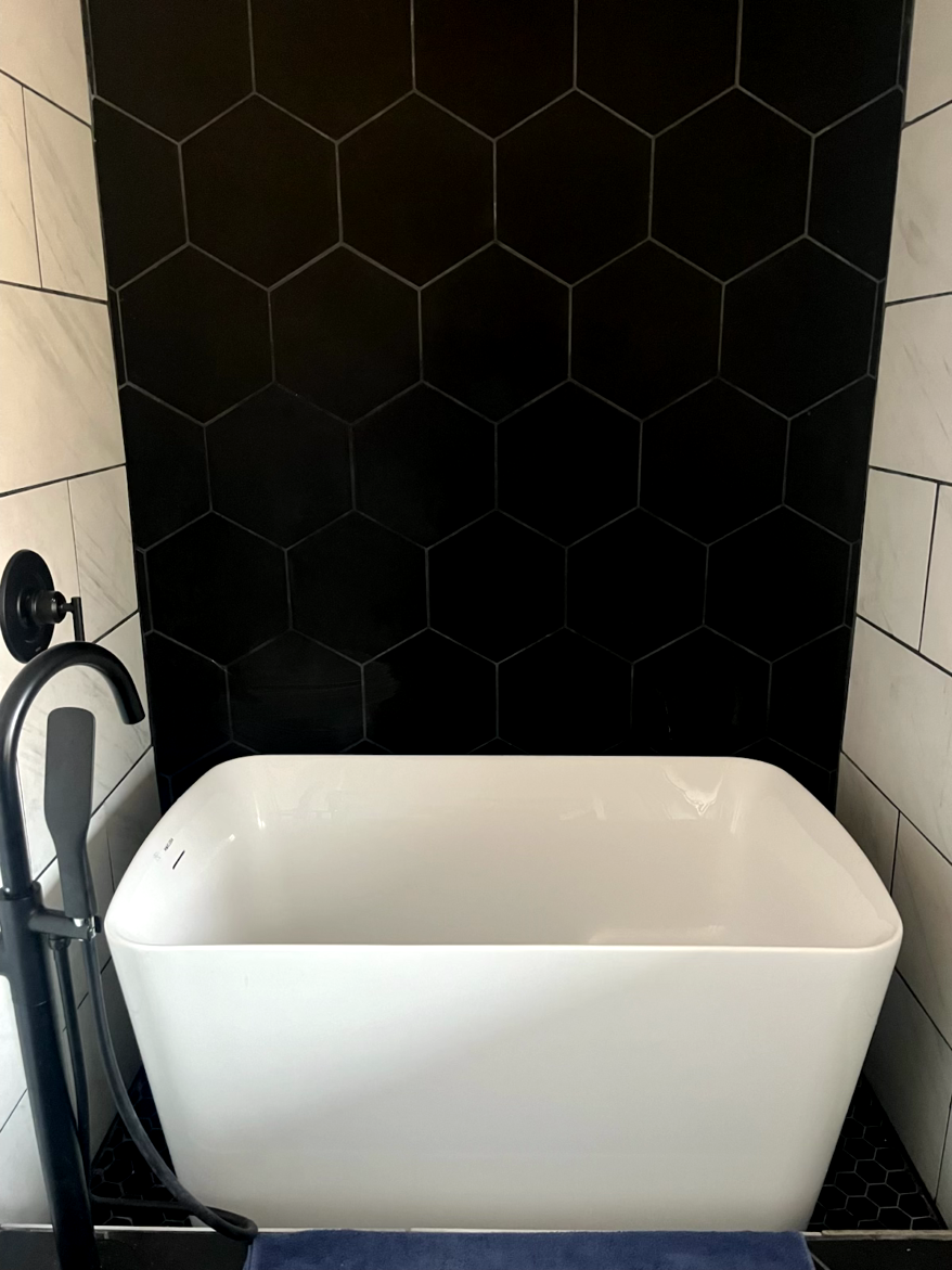 White freestanding bathtub against a black hexagon tile wall. Black faucet.
