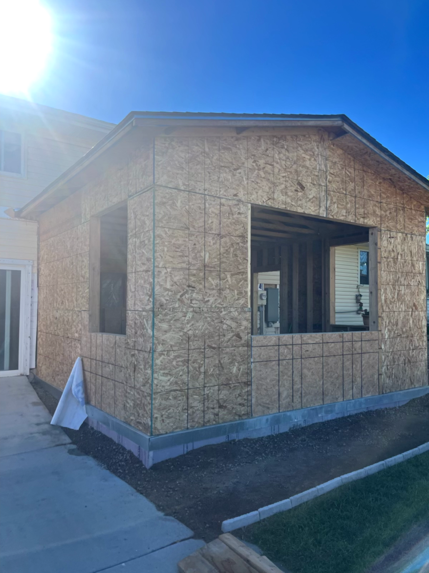 Construction of a house addition; exterior view showing plywood walls, window openings, and a concrete foundation.