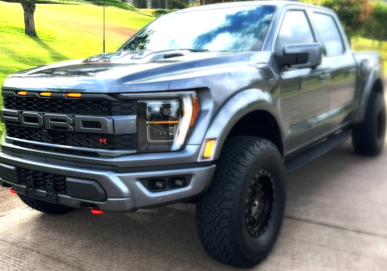 Gray Ford Raptor pickup truck parked on asphalt. Black wheels, off-road tires, and LED lights.