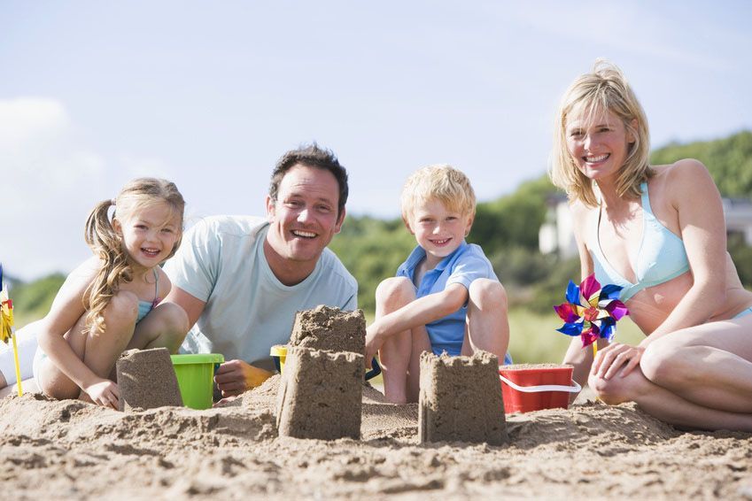 Familie bestehend aus Mutter, Vater, Tochter und Sohn sitzt am Strand vor Sandburg und lacht.