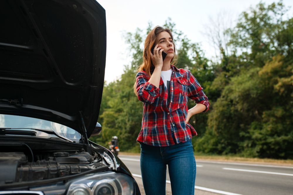 A woman is talking on a cell phone next to a broken down car.