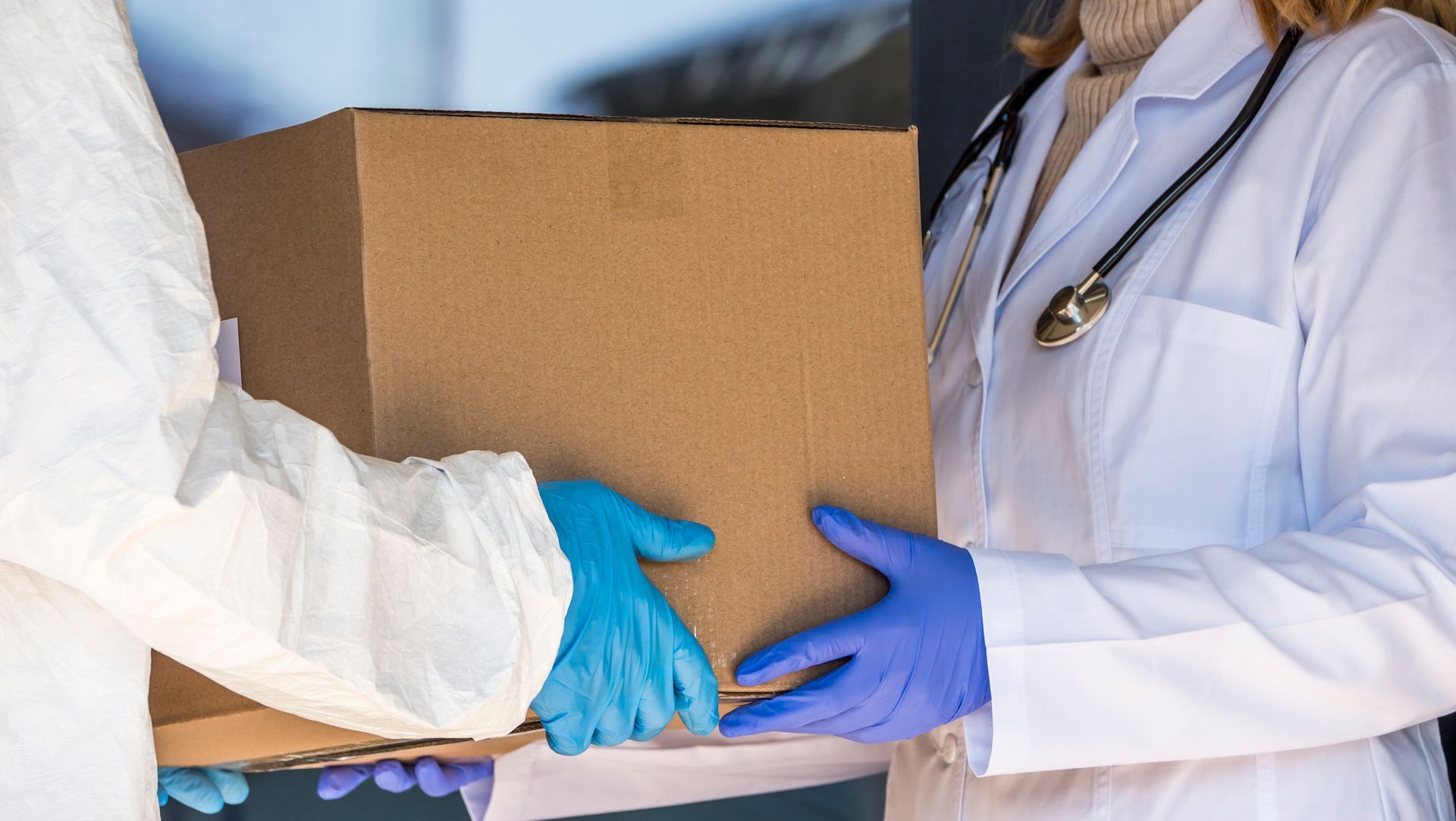 A doctor is handing a cardboard box to a person wearing blue gloves.