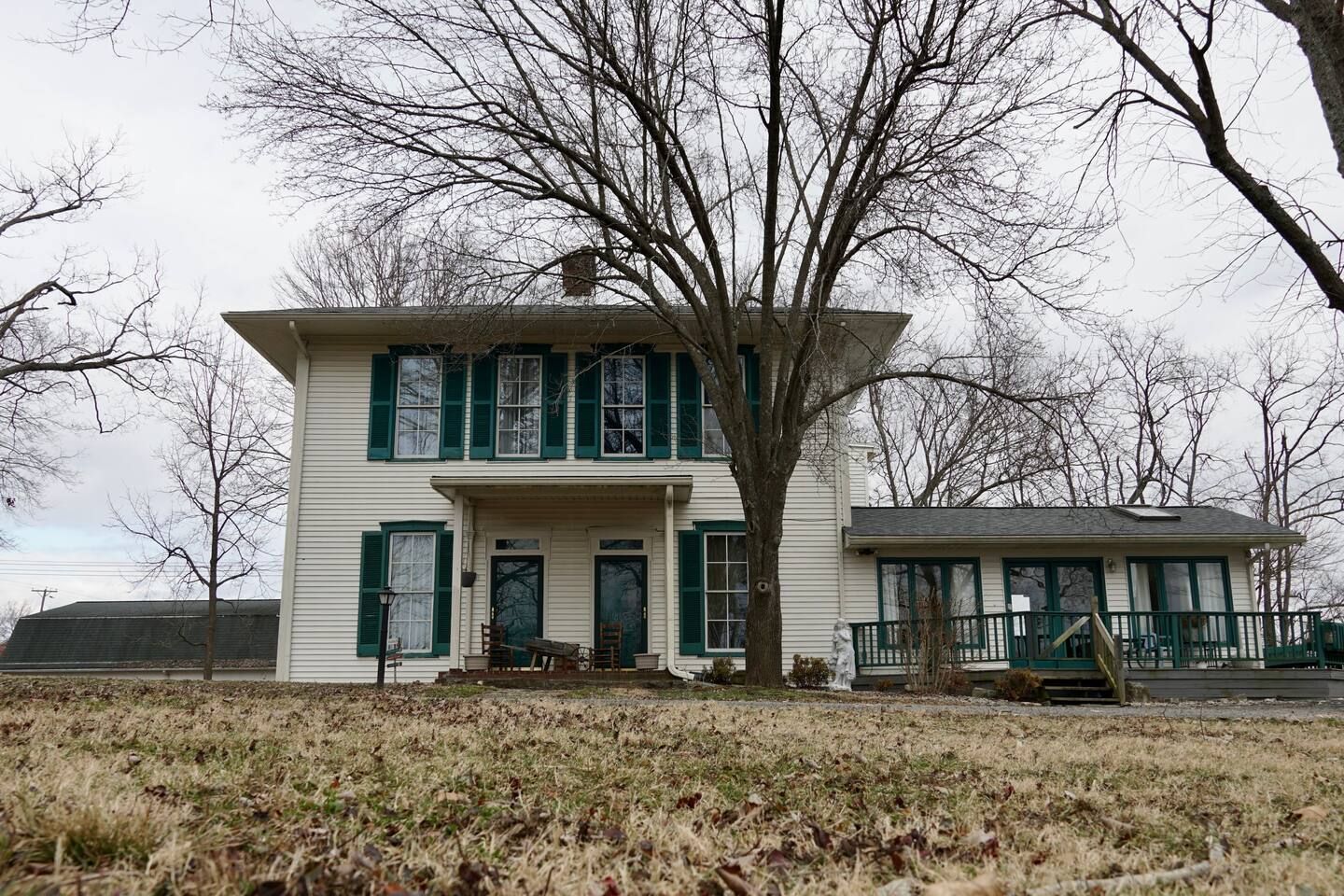 A large white house with green shutters is sitting in the middle of a grassy field.