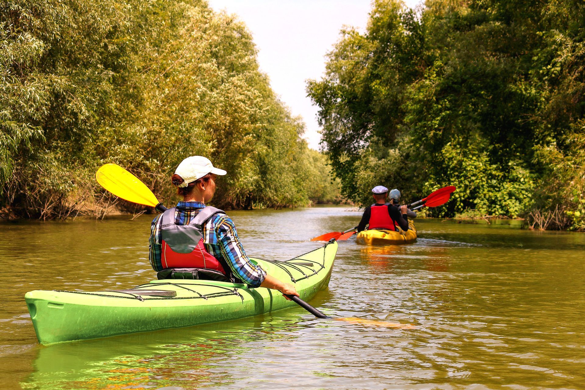 Two people kayaking on a green-tinted river bordered by trees. One person wears a white hat and paddles a green kayak.