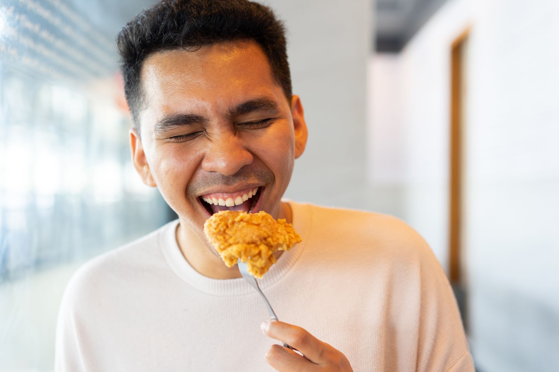 Man eating fried chicken, eyes closed in delight, indoors.