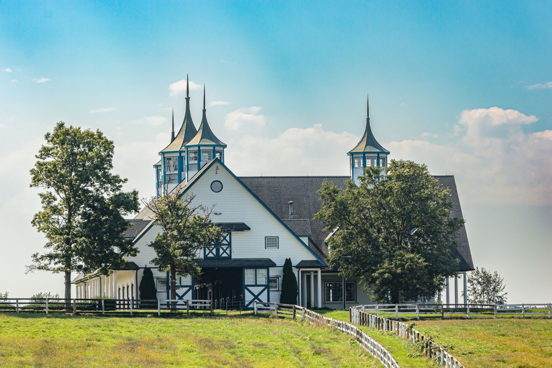 White barn with blue accents and decorative spires, surrounded by a fence and green field under a blue sky.