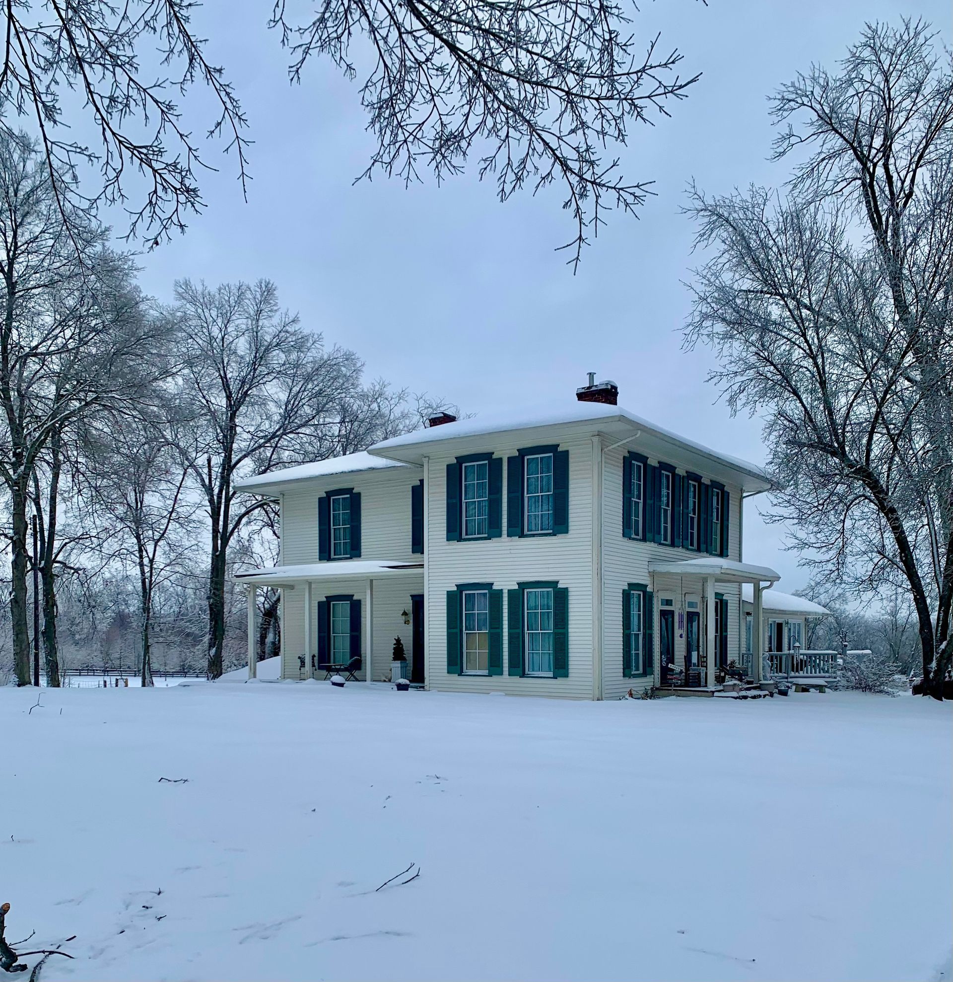 A white house with blue shutters is surrounded by snow and trees