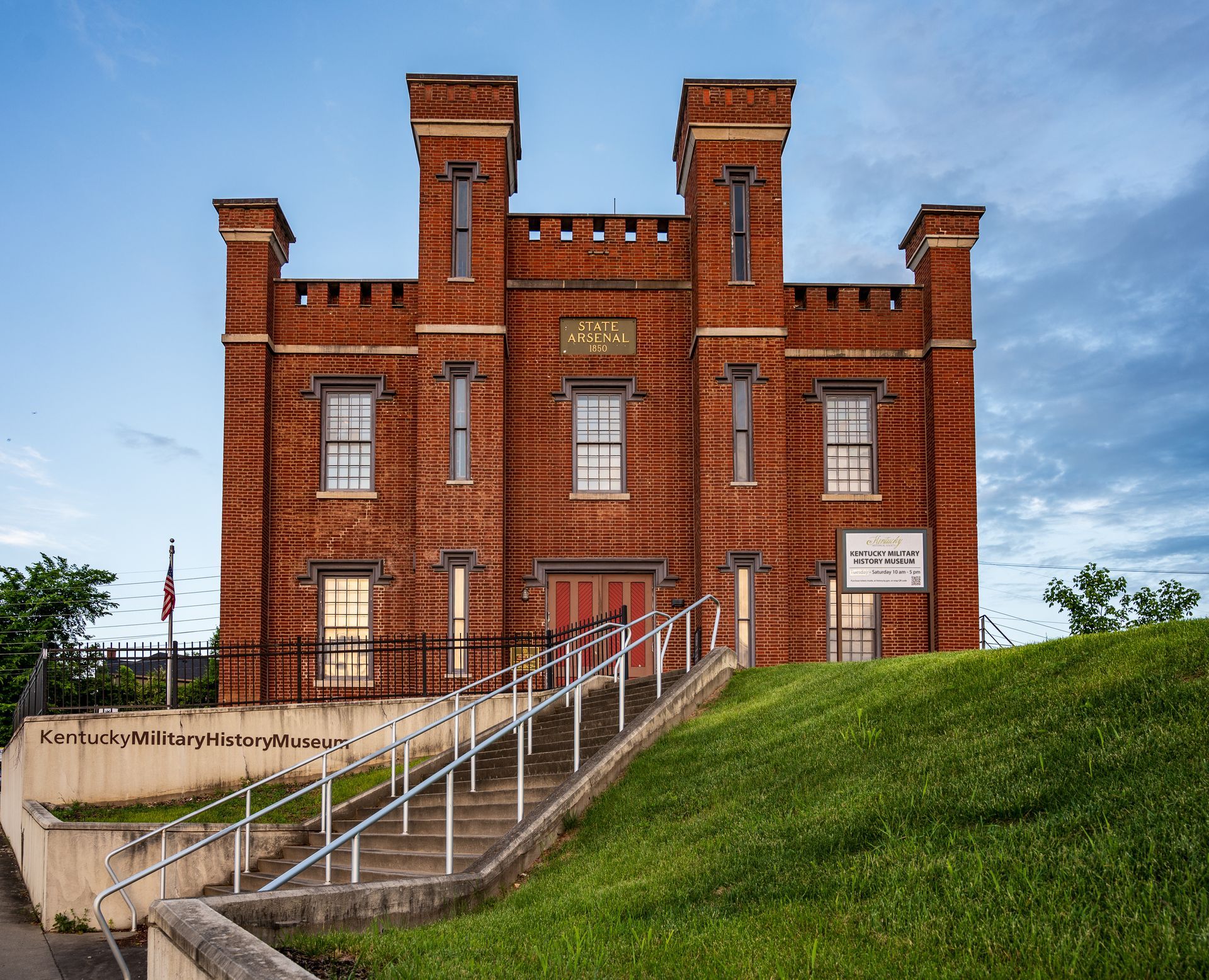 Red brick building with a central staircase, set on a grassy hill; American flag and sign present.