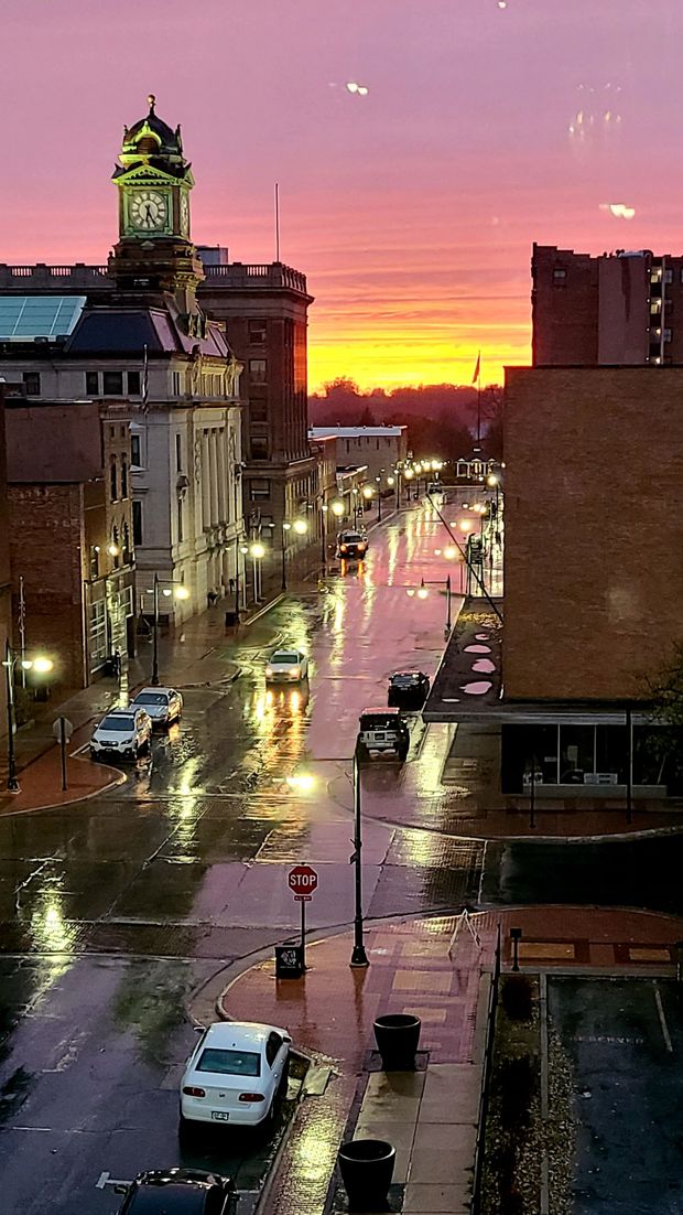 Wet city street at sunset, buildings with clock tower, cars driving, pink and orange sky.