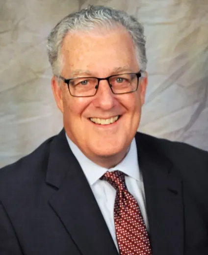 Man in glasses, suit, and patterned tie smiles; studio portrait.