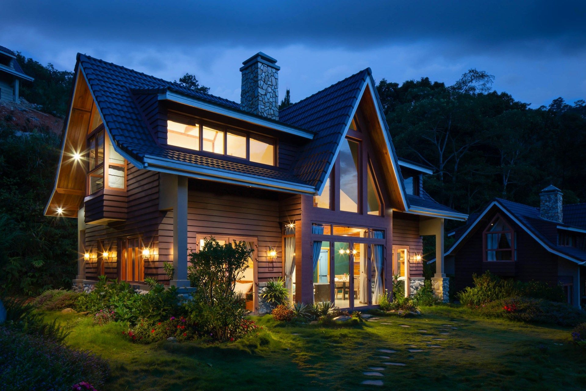 Lit-up wooden house with gabled roof at dusk, surrounded by greenery.