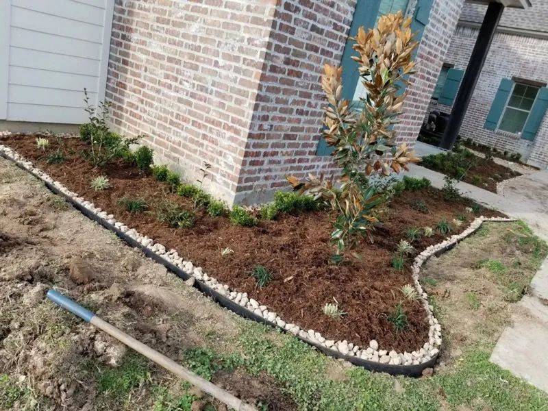 A newly landscaped flower bed in front of a brick house, mulched and edged with rocks.