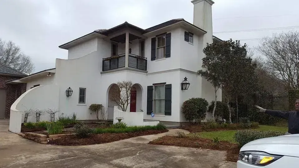 White stucco house with dark shutters, arched entryway, balcony, and a person pointing.