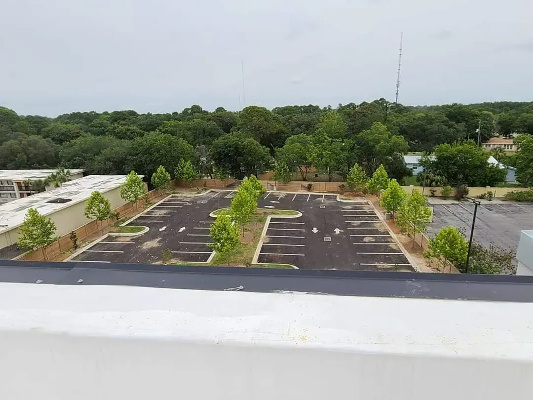 Overhead view of a parking lot with sparse trees in the center, surrounded by greenery and a cloudy sky.