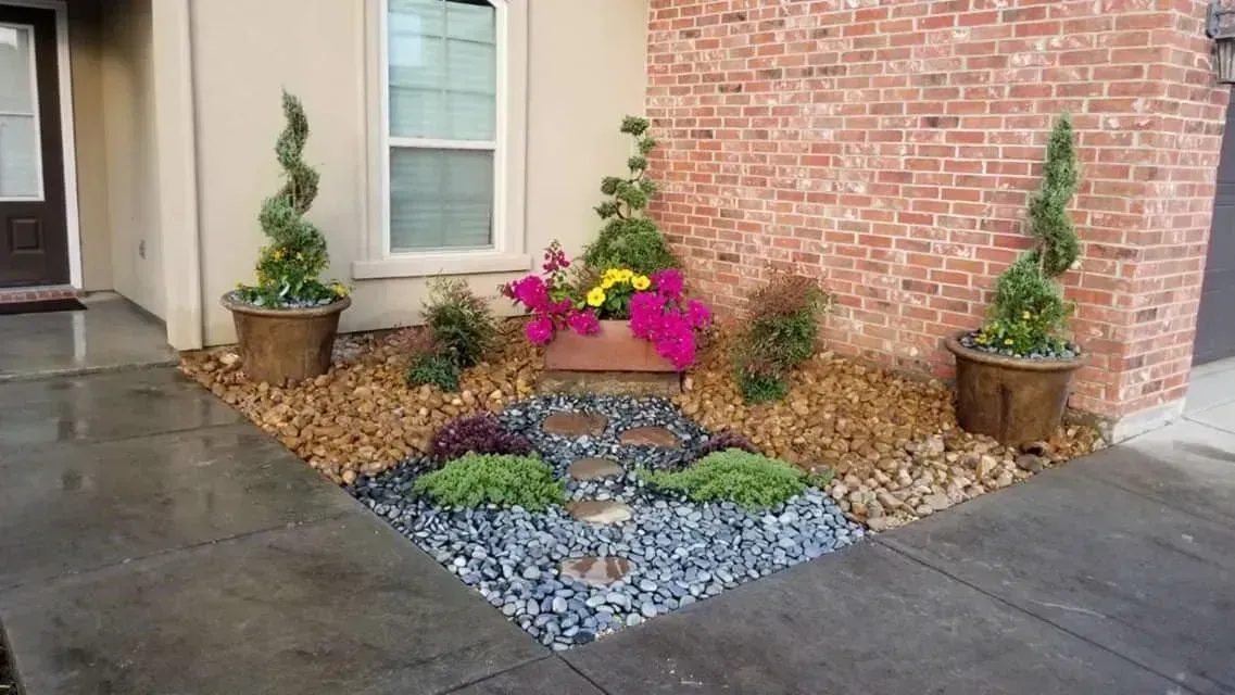 Landscaped corner of a building with potted plants, colorful flowers, and a rock path on a gray stone walkway.