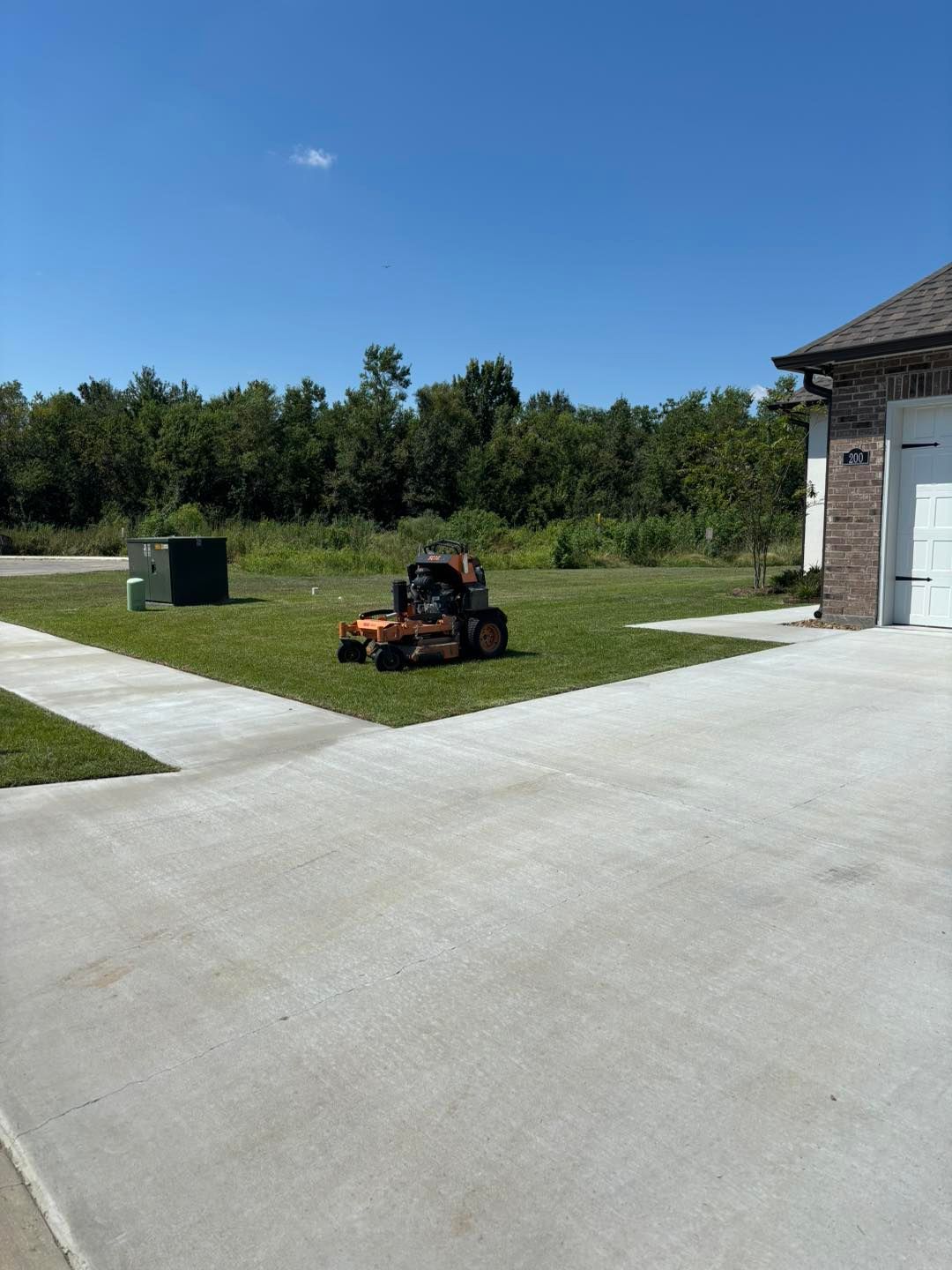 A lawn mower on a grassy yard, next to a concrete driveway and a house, under a blue sky.