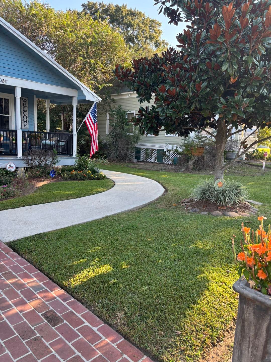 A paved path leads past a blue house with an American flag to another house under a tree.