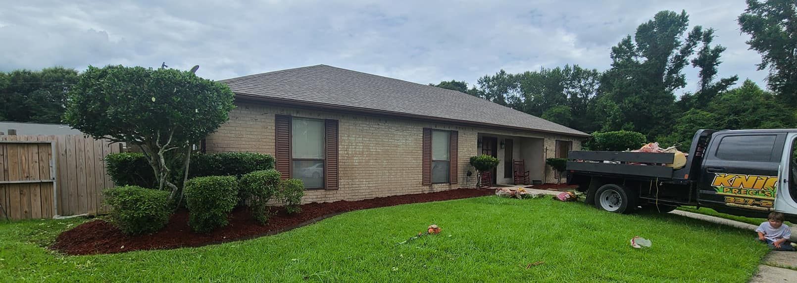 A house with brick facade and landscaped yard, a truck is parked in the yard. Overcast sky.