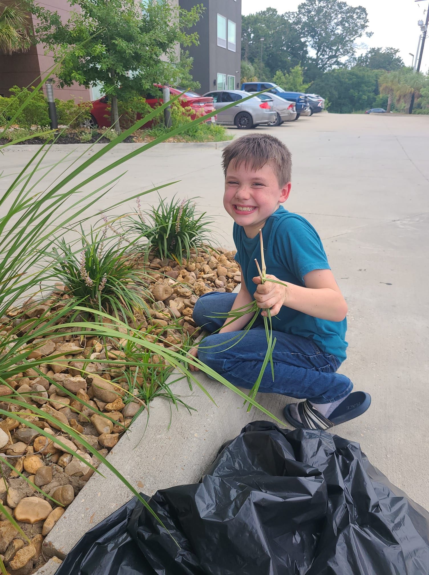 Boy kneeling, smiling, holding grass, near a rock garden and black trash bag.