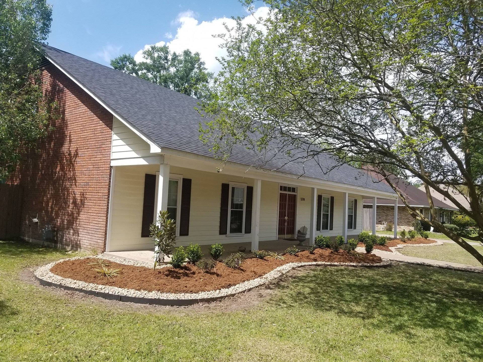 Beige house with black roof, brick chimney, porch, and landscaped flowerbed.