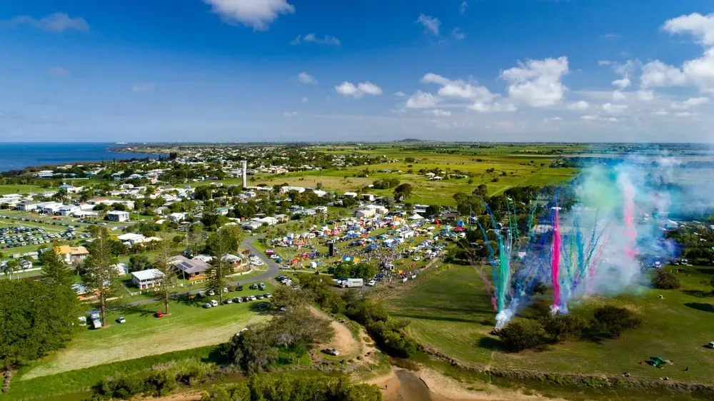 Aerial view of a town near a coastline with a gathering of people; colorful smoke plumes rise in the foreground. — Moore Park Beach Glass & Security in Burnett Heads, QLD