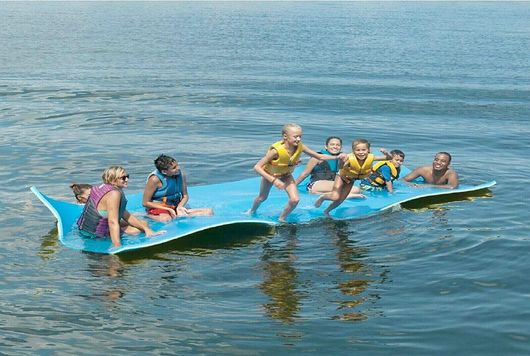 Six people on a blue floating platform on calm water, balancing together and smiling.