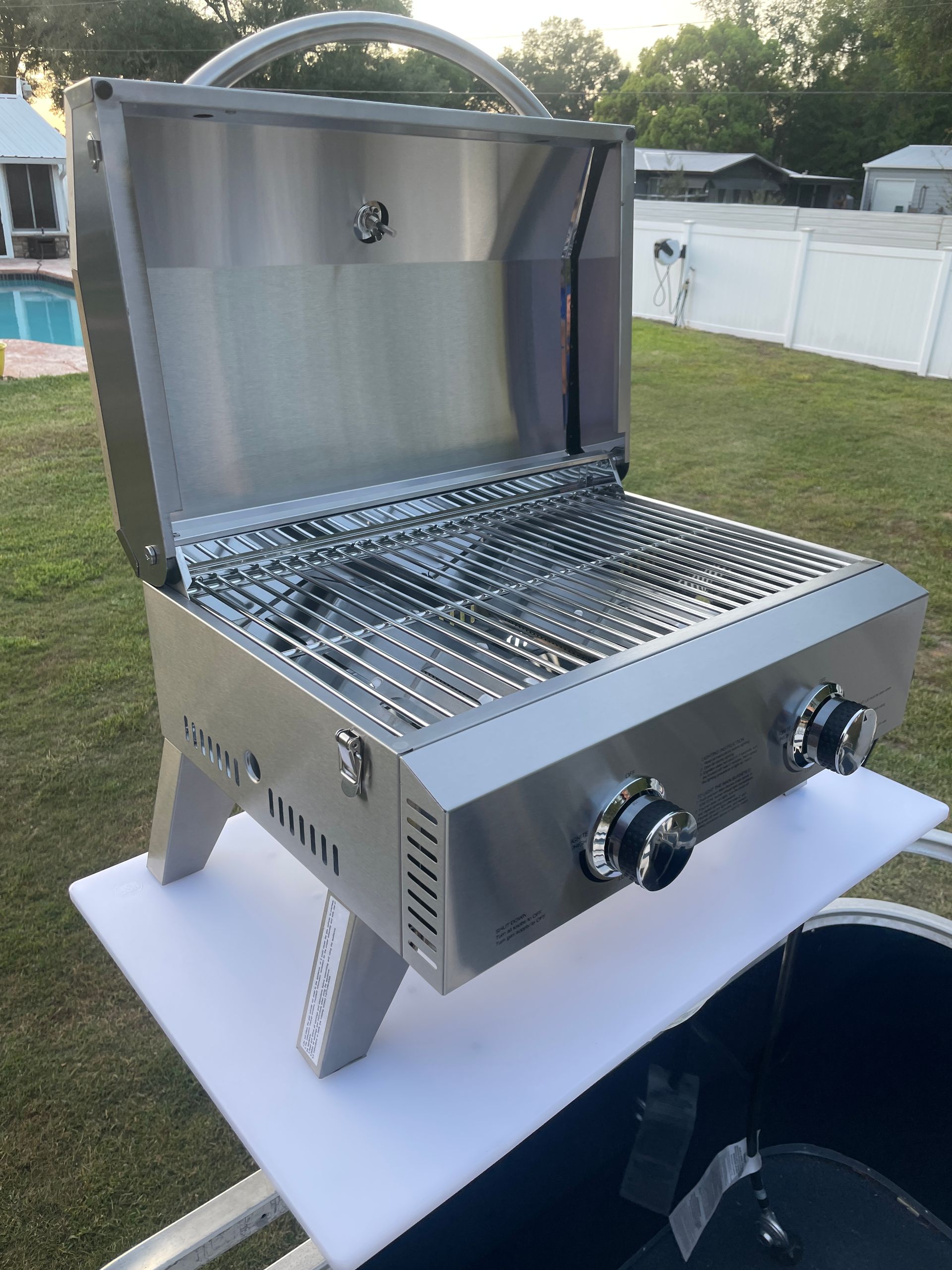 Stainless steel gas grill with lid open, sitting on a white table outdoors on grass.