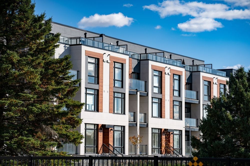 A large apartment building with a lot of windows is surrounded by trees and a fence.