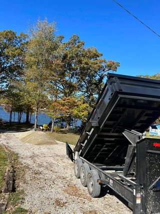 Black dump trailer tilted up, gravel driveway, lake and trees in background.