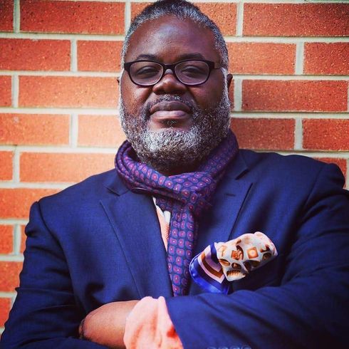 A man in a suit and scarf is standing in front of a brick wall