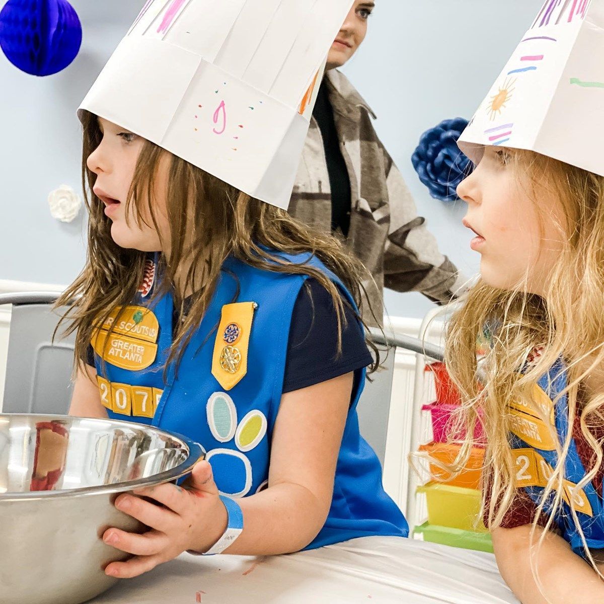 two girl scouts wearing paper hats are looking at a bowl