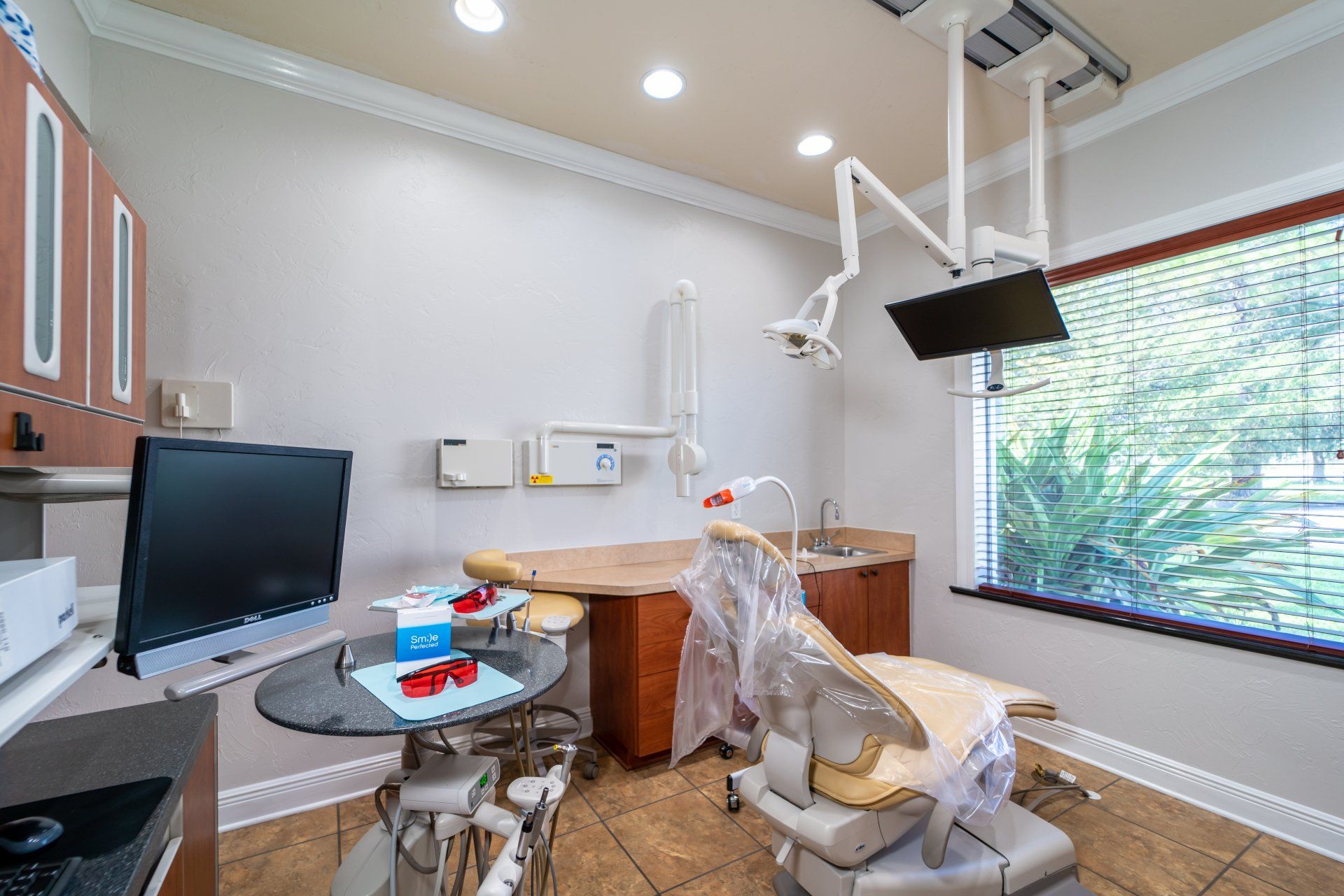 A dental office with a dental chair , desk , monitor and window.