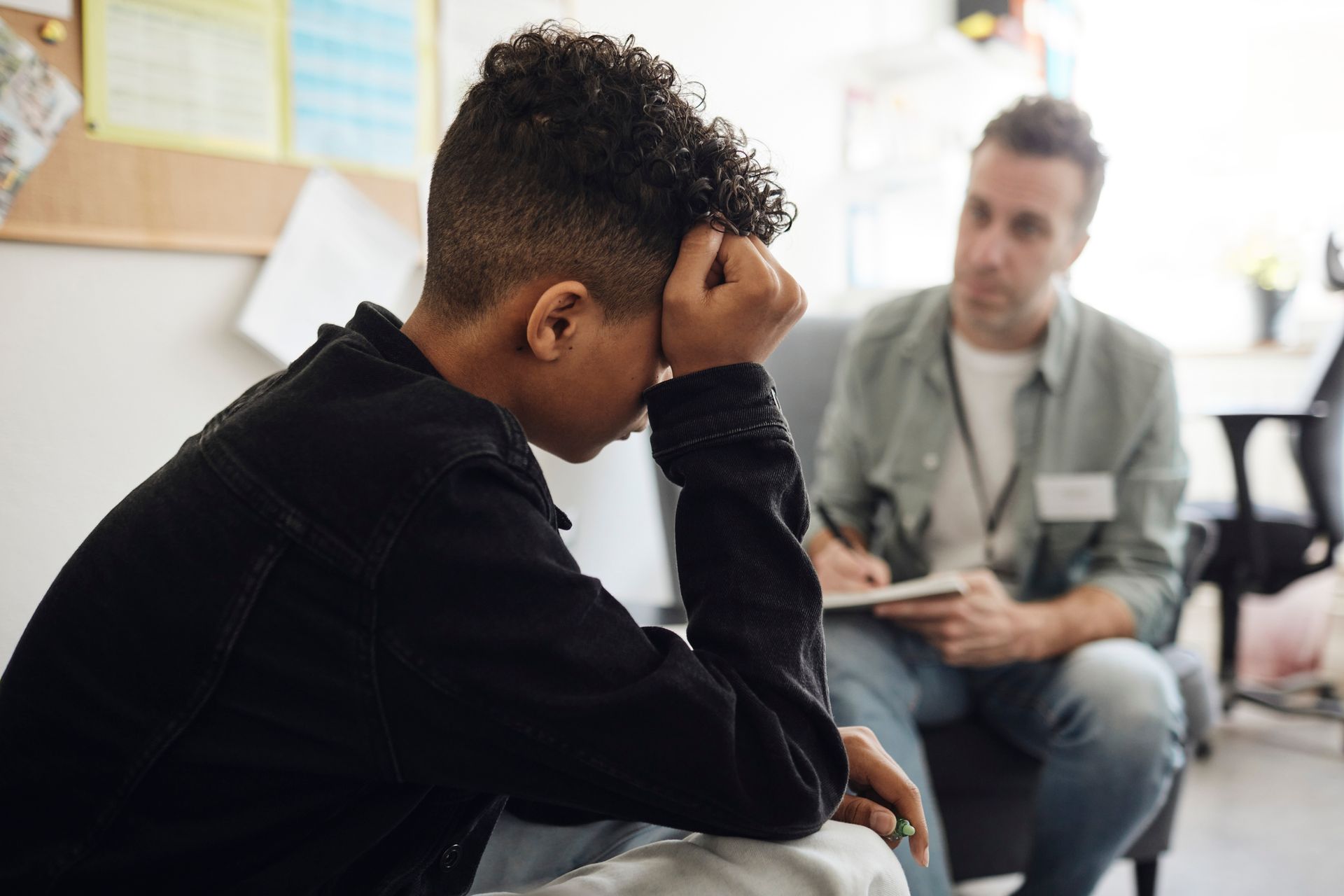 Young person with head in hands, sitting across from a person taking notes; an office setting.