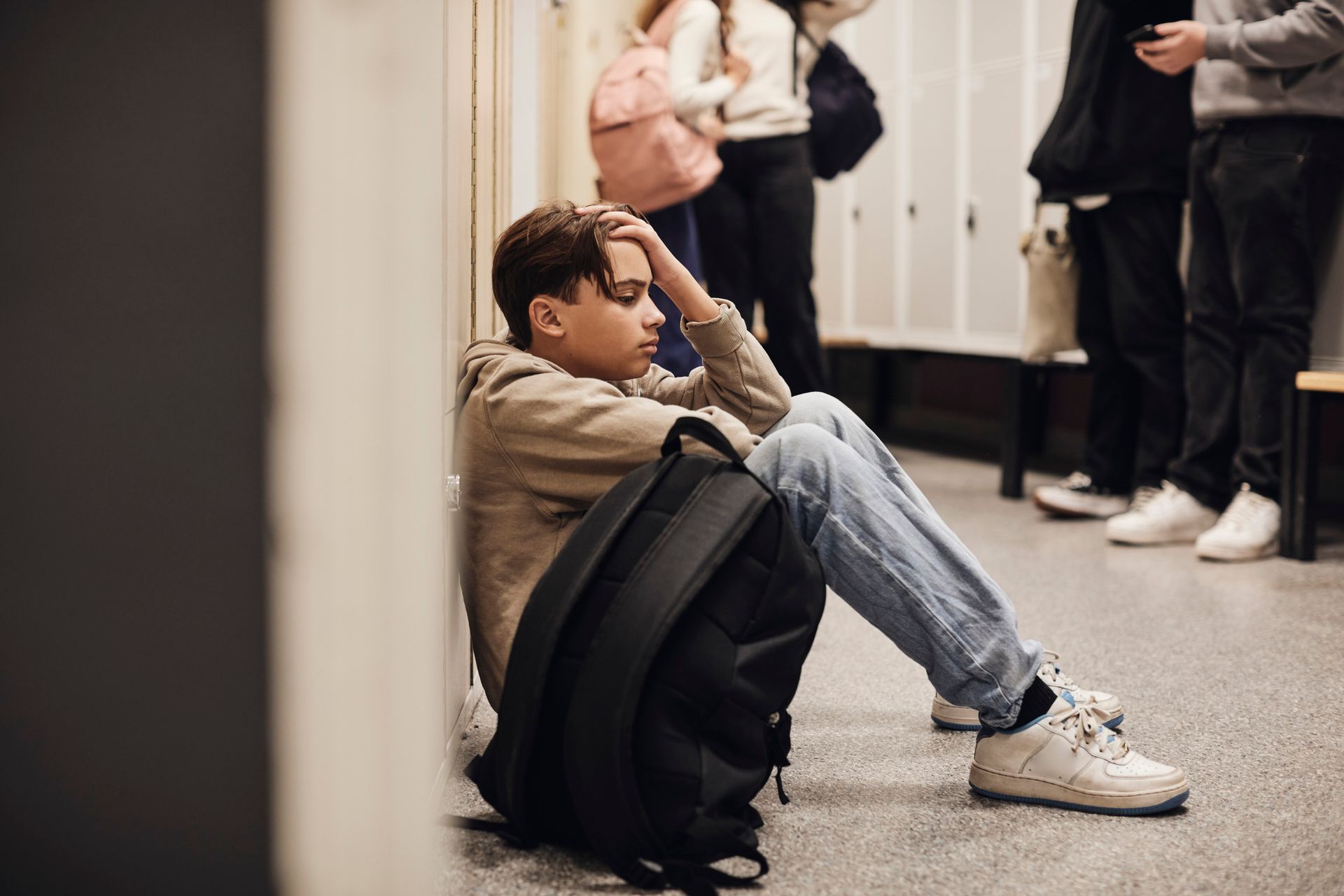 Boy sits against lockers, backpack beside him, head in hand, looking down, in school hallway.
