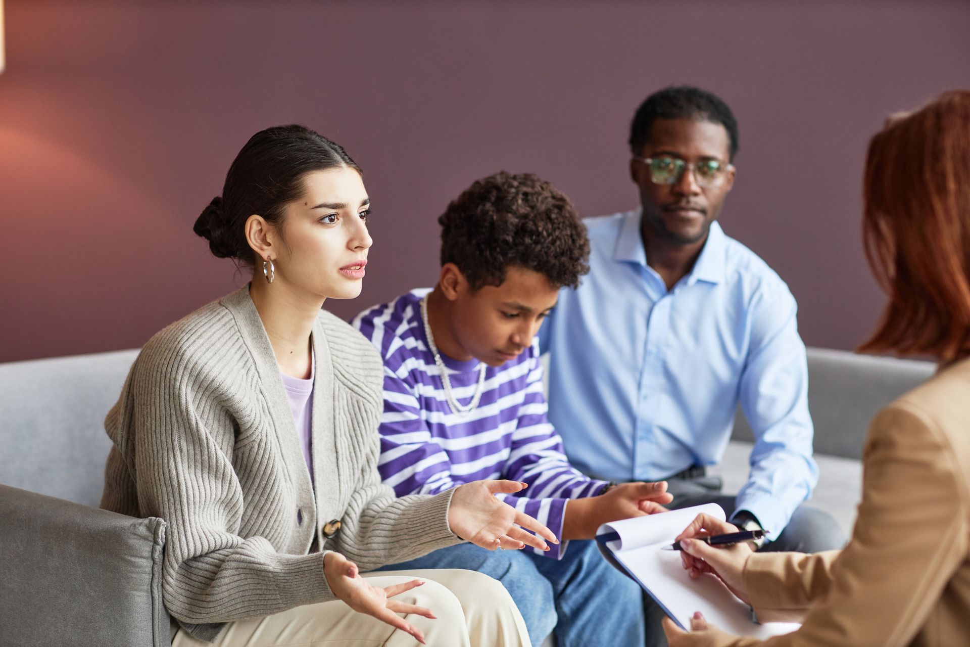 Family in therapy session; woman gestures, therapist takes notes.