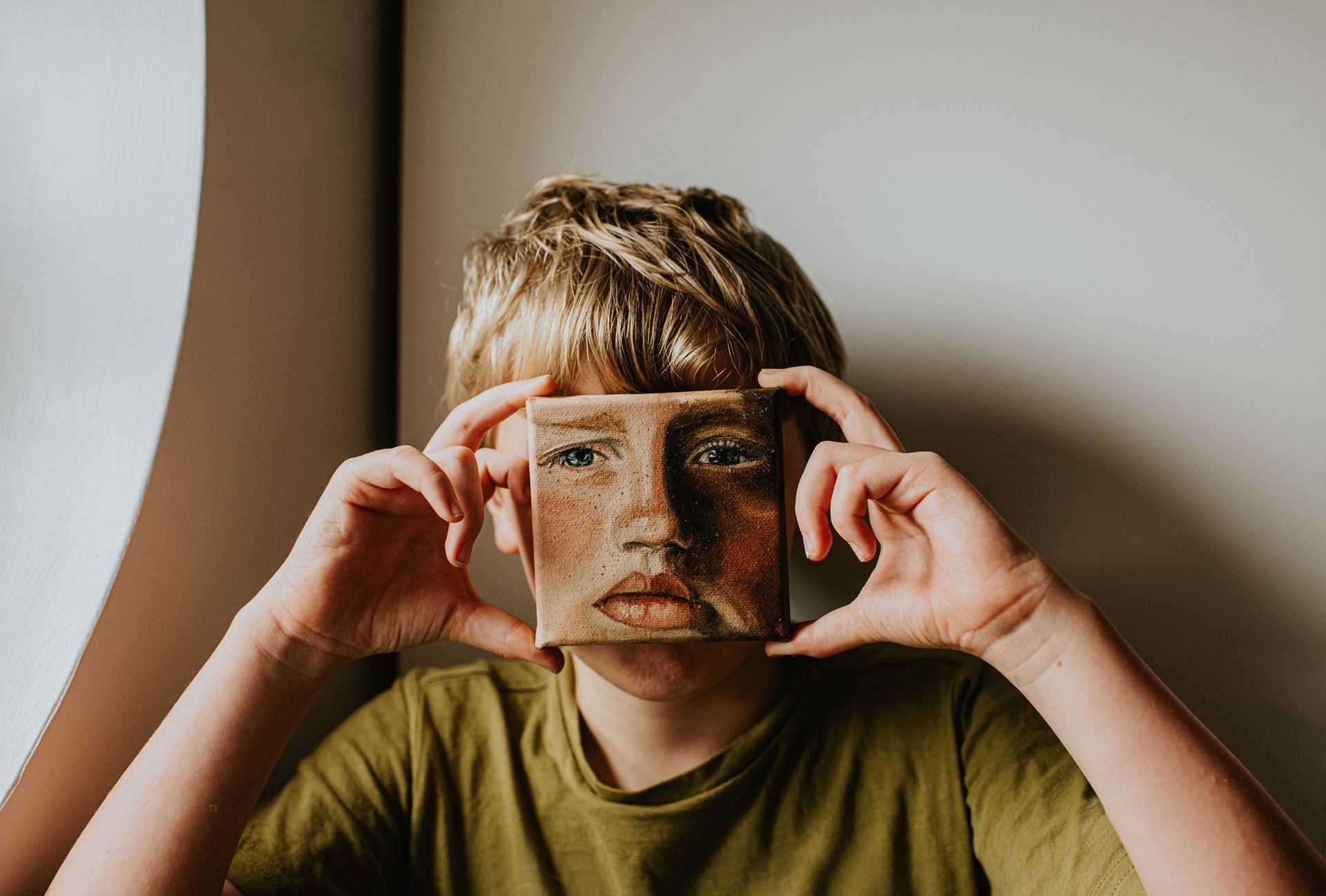 Person holding small square painting of a face in front of their own face.