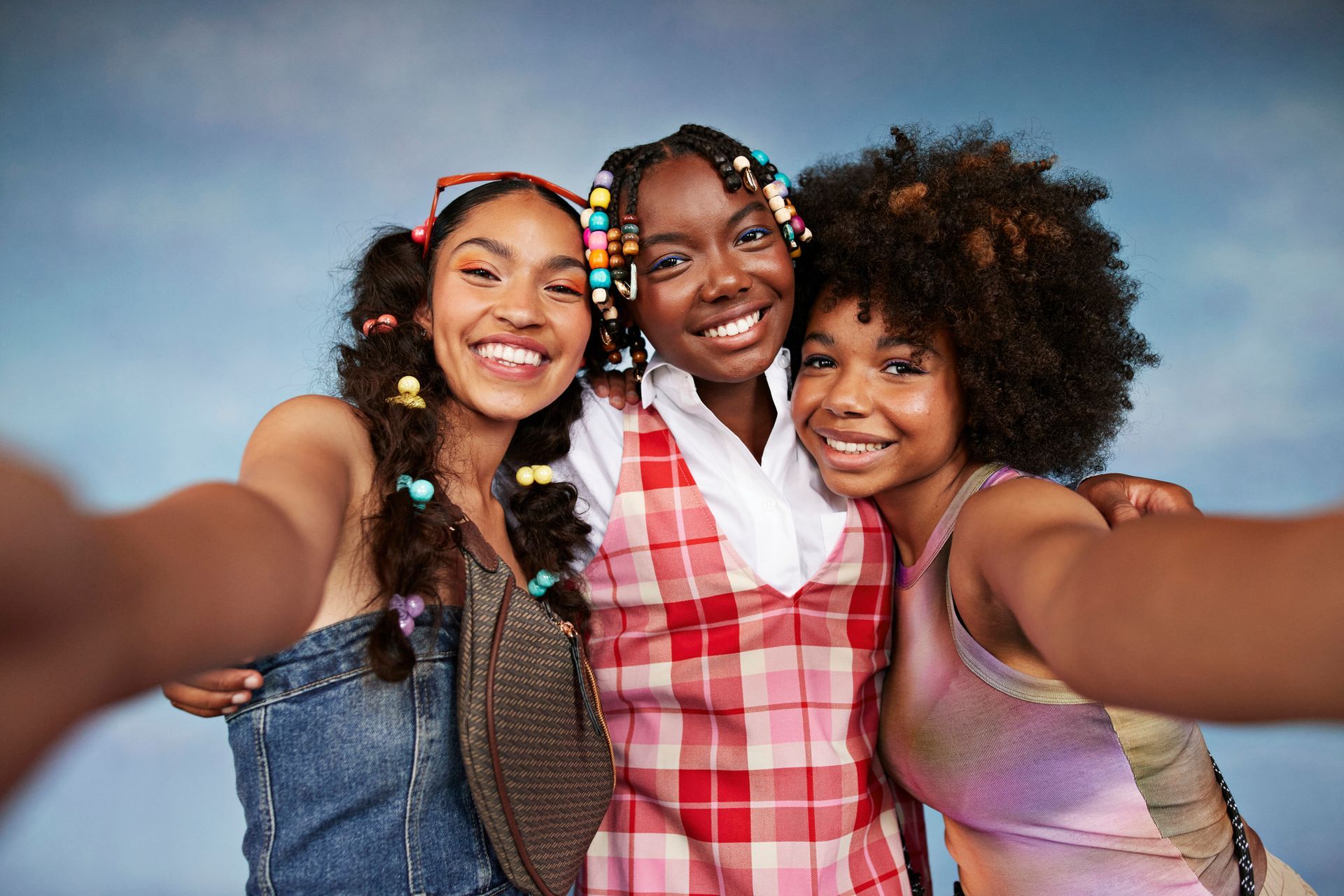 Three smiling people taking a selfie against a blue sky; one in a plaid dress, one with curly hair, and one with a denim vest.