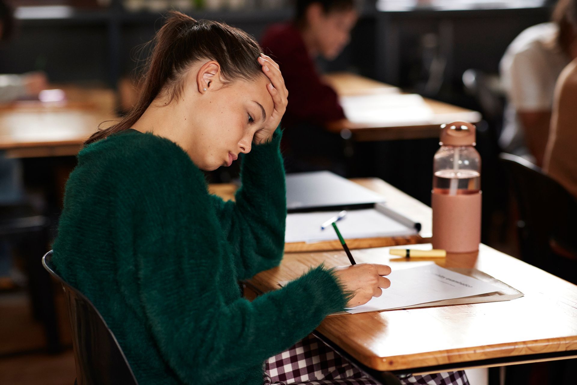 Woman with hand on forehead, writing at desk in classroom, looking stressed.