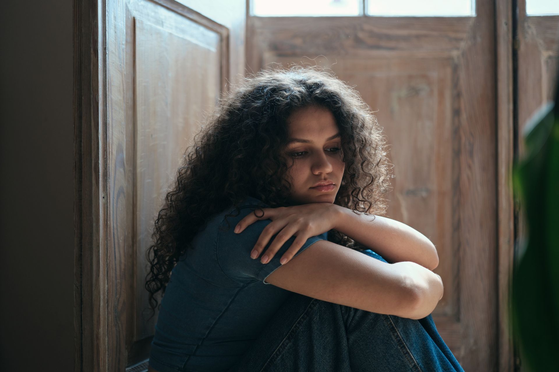 Woman with curly hair sits hugging knees, looking down near a wooden door.