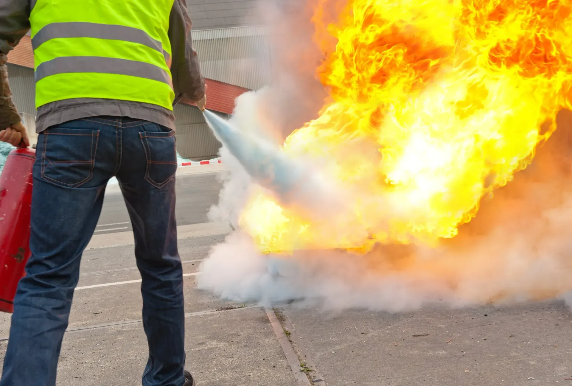A person wearing a high-visibility vest uses a red fire extinguisher to suppress bright orange flames outdoors.