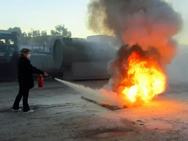 A person uses a fire extinguisher to combat a large, bright orange fire burning on the ground outdoors.